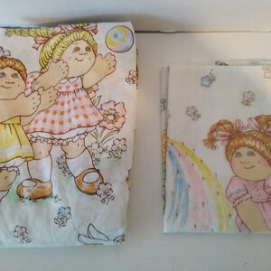 May include: Two folded white fabric sheets featuring Cabbage Patch Kids illustrations. The larger sheet displays two children in yellow and pink outfits with flowers and a ball. The smaller sheet shows a child in a pink dress near a rainbow.