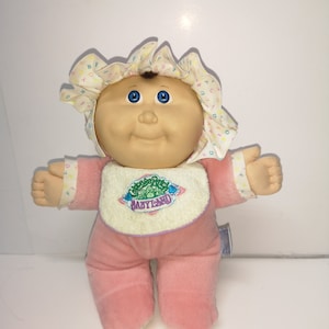 Peut inclure: Une poupée Cabbage Patch Kid portant une tenue rose avec un bavoir blanc qui dit "Cabbage Patch Kids BabyLand". La poupée a les yeux bleus et les cheveux peints en brun.