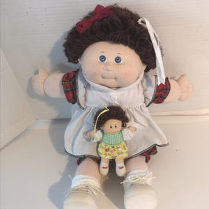 May include: A vintage doll with brown curly hair, blue eyes, and freckles, wearing a white dress with red plaid trim. The doll has a smaller doll attached. The doll is wearing white socks and shoes.