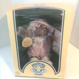 May include: A Cabbage Patch Kids Preemie doll in its original packaging. The doll has dark skin, wearing a pink and white striped outfit and bonnet. The box is yellow and green with the Cabbage Patch Kids logo and the text "Preemie!" and "I'm unique, I'm adoptable!"