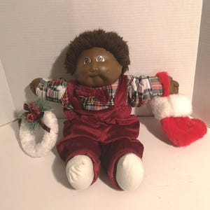 May include: A vintage doll with brown skin, wearing a red velvet outfit and a plaid shirt. The doll has brown hair and is holding a white wreath with red berries and a red and white Christmas stocking. A festive holiday decoration.