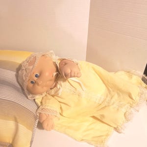 May include: A vintage baby doll with blue eyes, wearing a yellow dress with lace trim and a matching bonnet. The doll is lying on a striped pillow with a yellow blanket.