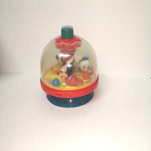 May include: A clear plastic spinning top toy with a red base and a green top. Inside the top are Mickey Mouse, Donald Duck, and Goofy figures.