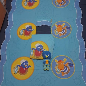 Peut inclure: Un ensemble de literie pour enfants bleu et jaune avec un personnage de pingouin et d'élan dessinés. L'ensemble comprend un drap-housse, un drap plat et une taie d'oreiller. La literie est décorée d'un thème nautique, y compris un volant, une ancre et des bulles.