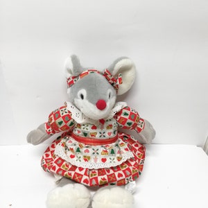 May include: A grey plush mouse wearing a red and white dress with a white lace trim. The dress has a pattern of houses, hearts, and Christmas trees. The mouse has a red nose and a white bow.