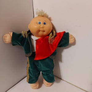 May include: A Cabbage Patch Kid doll wearing a green velvet suit with gold trim and a red sash. The doll has blonde hair and blue eyes.