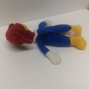 May include: A plush toy of Woody Woodpecker, a cartoon character. The toy is lying on its back with its head turned to the side. The toy is wearing a blue jumpsuit with white accents and yellow feet. The toy has a red head with a yellow beak.