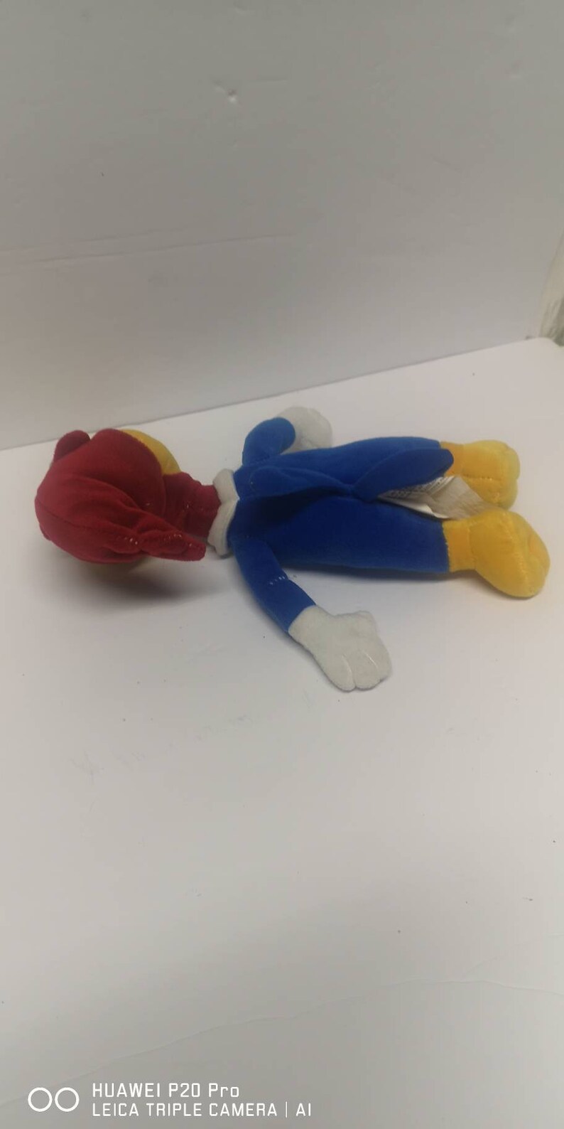 May include: A plush toy of Woody Woodpecker, a cartoon character. The toy is lying on its back with its head turned to the side. The toy is wearing a blue jumpsuit with white accents and yellow feet. The toy has a red head with a yellow beak.
