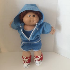 May include: A vintage doll with auburn hair, wearing a blue hooded tracksuit with a white zipper and matching shorts. The doll is wearing red and white crocheted sneakers with white laces. The doll is standing against a white background.