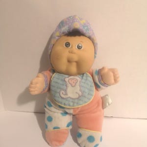 May include: A vintage Cabbage Patch Kids doll with a soft, plush body. The doll wears a pastel-colored bonnet and a bib with a monkey graphic. Its outfit includes pink and white polka-dot pants and matching sleeves.