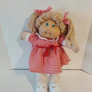 Peut inclure: Une poupée aux cheveux en laine blonde coiffés en couettes avec des nœuds à carreaux rouges et blancs. La poupée porte une robe vichy rouge et blanche avec un col en dentelle blanche, un ruban rouge à la taille, des collants en dentelle et des chaussures blanches.