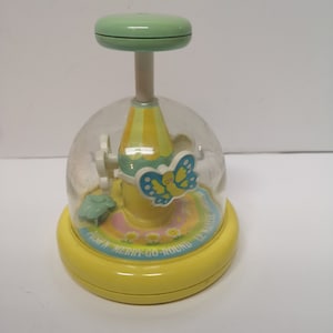 May include: A yellow plastic toy merry-go-round with a clear dome and a green top. The merry-go-round has a yellow butterfly with blue wings and a yellow and white striped tent. The text on the base reads "Push 'n Merry-Go-Round The Magic"
