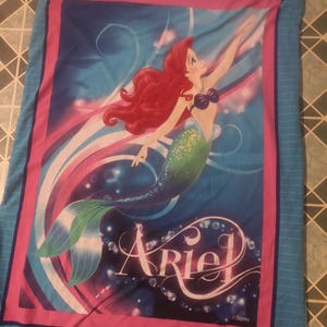 Puede incluir: Una manta azul con una vibrante ilustraci&oacute;n de Ariel, la Sirenita, con cabello rojo y cola verde. El dise&ntilde;o incluye el nombre "Ariel" en una fuente estilizada, sobre un fondo de colores arremolinados y burbujas.