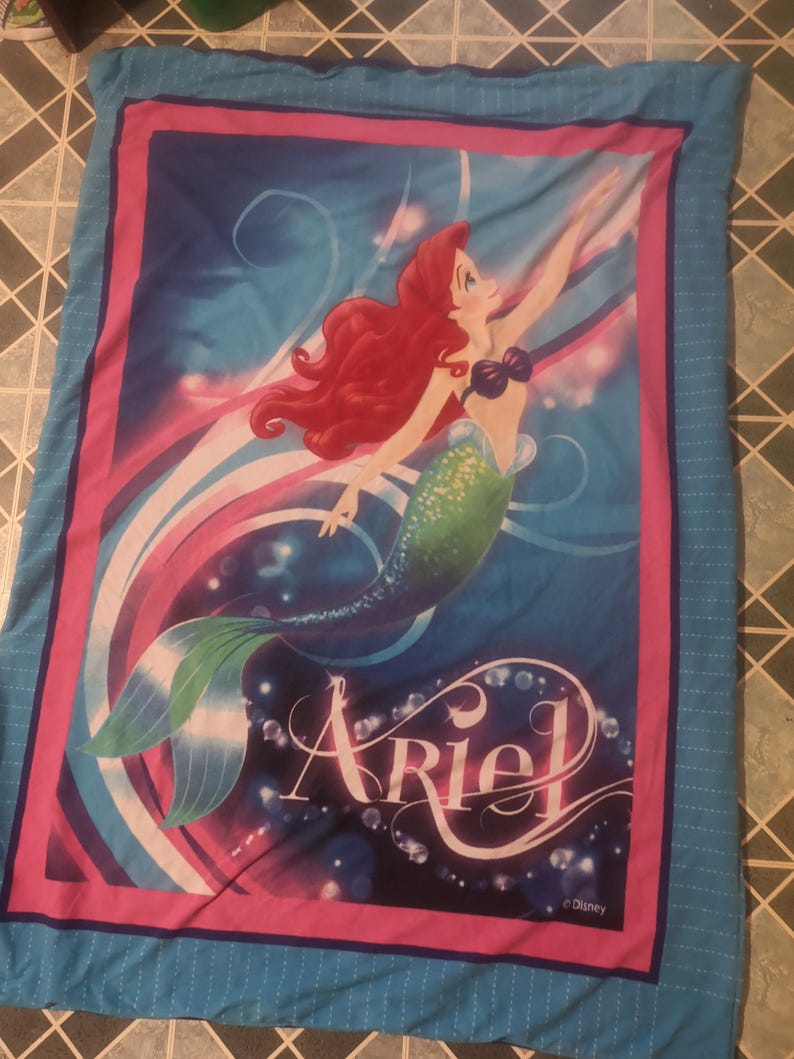 Puede incluir: Una manta azul con una vibrante ilustraci&oacute;n de Ariel, la Sirenita, con cabello rojo y cola verde. El dise&ntilde;o incluye el nombre "Ariel" en una fuente estilizada, sobre un fondo de colores arremolinados y burbujas.