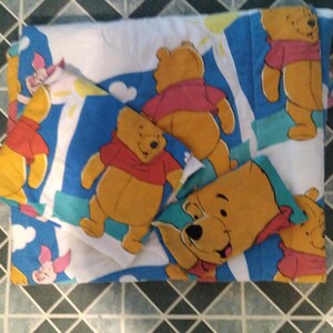 May include: A quilted bedding set featuring a repeating pattern of Winnie the Pooh and Piglet on a white background with blue accents. The design includes multiple illustrations of the yellow bear in a red shirt and the small pink pig character.