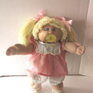 May include: A doll with blonde yarn hair styled in pigtails with pink ribbons, blue eyes, and a gold pacifier in its mouth. The doll wears a pink and white gingham dress with a white collar and lace trim, white bloomers, and white shoes.