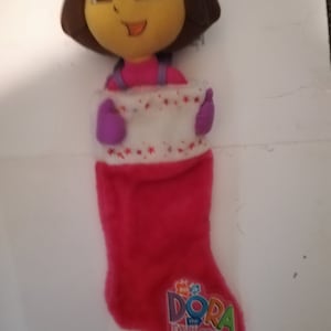 May include: A red and white Christmas stocking with a plush Dora the Explorer character peeking out of the top. The stocking has a white cuff with red stars and the words "Dora the Explorer" in colorful letters.