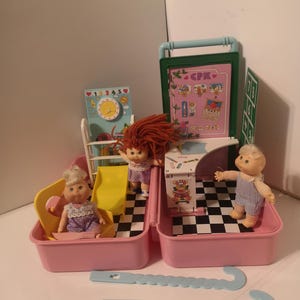 May include: A pink plastic carrying case with a built-in playset for dolls. The case has a chequered black and white floor, a yellow slide, a desk with a chair, and a chalkboard with the text "CPK". There are three dolls included, two with blonde hair and one with red hair.