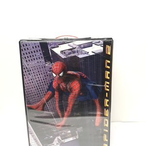 May include: A black DVD case with a red handle. The front of the case features a graphic of Spider-Man swinging through a city skyline. The text "Spider-Man 2" is printed in gold on the right side of the case.