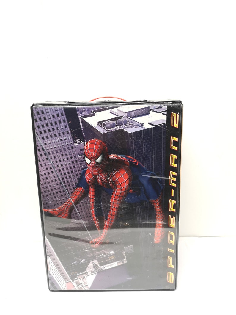 May include: A black DVD case with a red handle. The front of the case features a graphic of Spider-Man swinging through a city skyline. The text "Spider-Man 2" is printed in gold on the right side of the case.