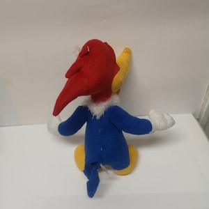 Puede incluir: Un juguete de peluche de Woody Woodpecker, un personaje de dibujos animados. El juguete es rojo, amarillo y azul. El juguete est&aacute; sentado sobre una superficie blanca.