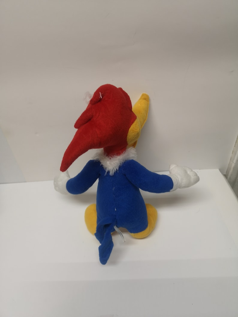 Puede incluir: Un juguete de peluche de Woody Woodpecker, un personaje de dibujos animados. El juguete es rojo, amarillo y azul. El juguete est&aacute; sentado sobre una superficie blanca.