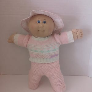 May include: A vintage doll with a soft, vinyl face and blue eyes, wearing a pink knitted outfit and a matching hat. The doll's sweater has a pastel floral design. The doll's arms are outstretched, and it is standing on a white surface.