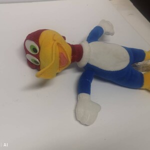 May include: A plush toy of Woody Woodpecker, a cartoon character. The toy is lying on its back with its arms outstretched. The toy is yellow, red, blue, and white.