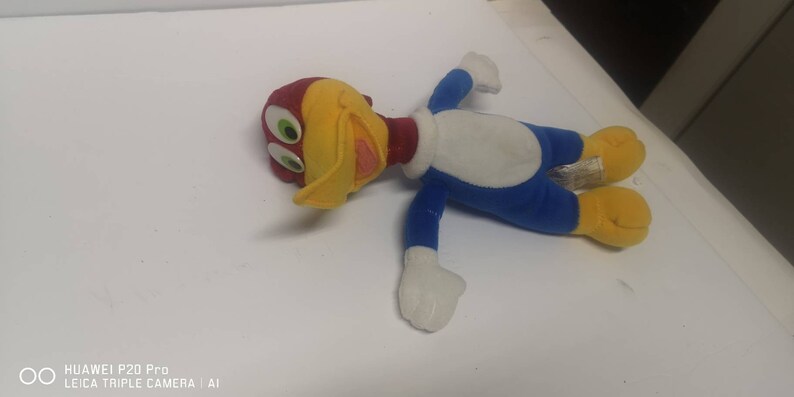 May include: A plush toy of Woody Woodpecker, a cartoon character. The toy is lying on its back with its arms outstretched. The toy is yellow, red, blue, and white.