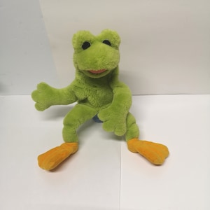 May include: Green plush frog puppet with orange feet. The frog has black eyes and a wide, toothy smile.