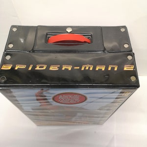 May include: A black plastic carrying case with a red handle. The case has the words "Spider-Man 2" printed in gold on the front. The case has a sticker on the side that says "2 Spider-Man Movie Action Figures Inside!" and "2 figurines d'action Spider-Man &agrave; l'int&eacute;rieur."