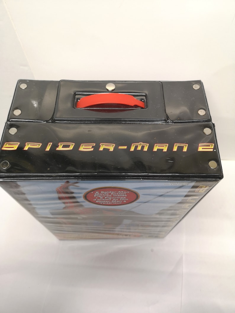 May include: A black plastic carrying case with a red handle. The case has the words "Spider-Man 2" printed in gold on the front. The case has a sticker on the side that says "2 Spider-Man Movie Action Figures Inside!" and "2 figurines d'action Spider-Man &agrave; l'int&eacute;rieur."