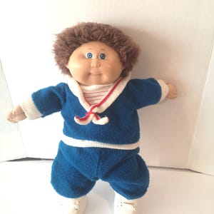 May include: A doll with brown yarn hair and blue eyes, wearing a blue fleece sailor-style outfit with a white collar, red trim, and white ribbed cuffs. The doll has light skin, a round face, and is wearing white shoes with white laces.