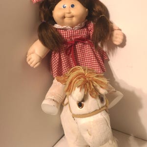 May include: A Cabbage Patch Kid doll with brown hair and a red and white gingham dress riding a white plush horse with brown mane and tail.