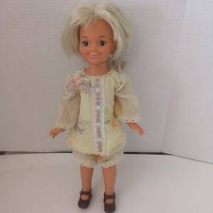 May include: A vintage doll with blonde hair, dressed in a yellow floral print outfit with lace detailing. The doll is wearing brown shoes and is standing. The outfit consists of a top and shorts.