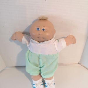 May include: A vintage Cabbage Patch Kid doll with a bald head and blue eyes. The doll is wearing a white shirt, mint green shorts, and white sneakers with blue stripes. The doll's arms are outstretched.