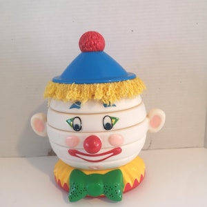 May include: A vintage clown toy featuring a blue hat, yellow fringe, and a red pom-pom. The clown has a white face with painted eyes, a red nose, and a red smile. A green bow tie and yellow collar complete the design.