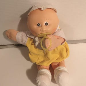 May include: A vintage Cabbage Patch Kids doll with a peach-coloured face, dark eyes, and a dummy. The doll wears a white bonnet, a white shirt, and a yellow corduroy romper. White socks cover the doll's feet.