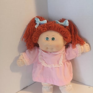 May include: A vintage Cabbage Patch Kid doll with red yarn hair styled in pigtails, adorned with light blue bows. The doll is dressed in a pink dress with lace trim and matching socks. The doll has green eyes and a soft, rounded face.