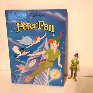 May include: A blue book with the title "Walt Disney's Peter Pan" on the cover. The cover features an illustration of Peter Pan flying with Tinkerbell, Wendy, and the Lost Boys. A green Peter Pan action figure is standing to the right of the book.