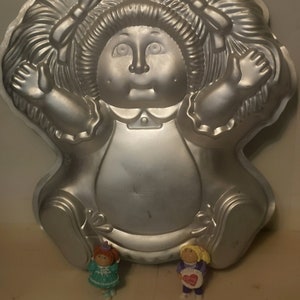 May include: A silver metal cake pan shaped like a Cabbage Patch Kid doll. The doll has a red bow in her hair and is wearing a dress.