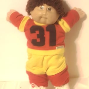 May include: A doll with brown yarn hair and a round face wearing a red and yellow long-sleeved shirt with the number 31 printed in black on the chest. The doll is dressed in matching red and yellow pants and white shoes with laces.