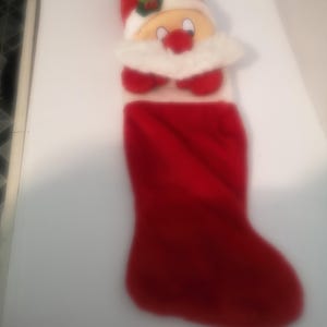 May include: A festive red Christmas stocking featuring a Santa Claus design. The top section showcases Santa's face with a red nose, white beard, and a red hat with white trim. The stocking is made of a soft, plush material.