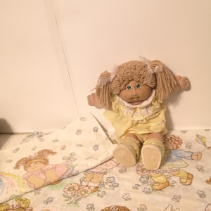 May include: A Cabbage Patch Kid doll with blonde hair and a yellow dress is sitting on a white and blue bedding set with a Cabbage Patch Kid print. The bedding set features a floral pattern and images of Cabbage Patch Kids playing.