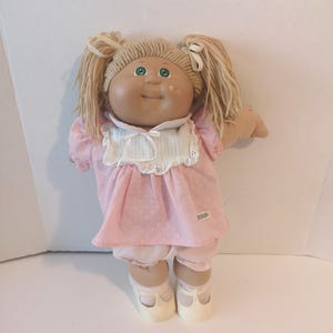 Peut inclure: Une poupée avec des cheveux en laine blonde coiffés en deux couettes, des yeux verts et un visage rond. La poupée porte une robe rose à pois blancs, un col blanc à rayures bleues et une bordure en dentelle. Elle porte aussi un bloomer rose assorti.
