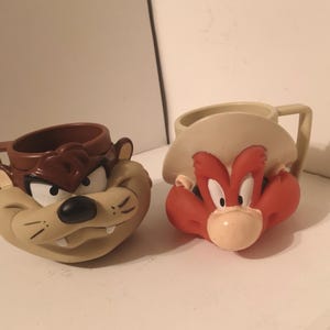 May include: Two ceramic mugs featuring cartoon characters from the Looney Tunes franchise. The mug on the left features a brown and tan Tasmanian Devil with a menacing expression. The mug on the right features a red and tan Yosemite Sam with a cowboy hat and a stern expression.