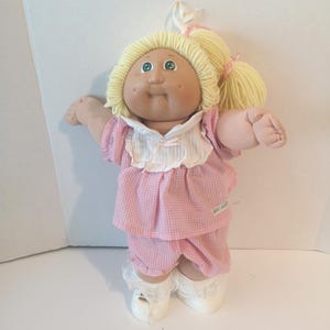Peut inclure: Une poupée en tissu avec des cheveux en laine coiffés en deux queues de cheval, des yeux verts et des joues avec des fossettes. La poupée porte un haut à carreaux vichy rose et blanc avec un col blanc, un short assorti, des chaussettes en dentelle.