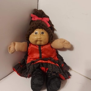 May include: A Cabbage Patch Kid doll with brown hair and a red bow. The doll is wearing a red dress with black lace trim and black shoes.