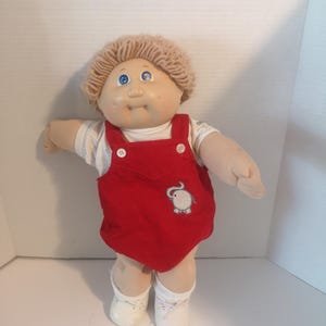May include: A vintage Cabbage Patch Kid doll with fair skin, blue eyes, and a light brown yarn hairstyle. The doll wears a red corduroy romper with an embroidered elephant and white shoes.