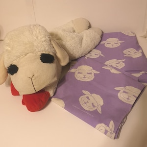 May include: A white plush sheep toy with a red nose lying on a purple sheet with a white sheep pattern.